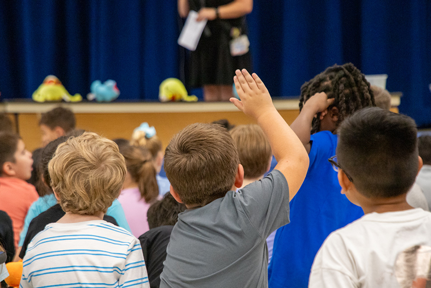 A boy in a gray shirt raises his hand during the assembly