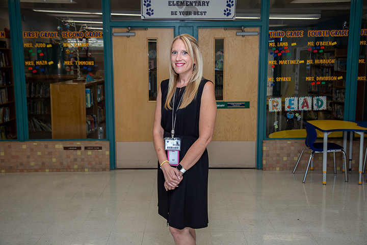 A woman with blonde hair wearing a black dress stands in the school hallway in front of the library